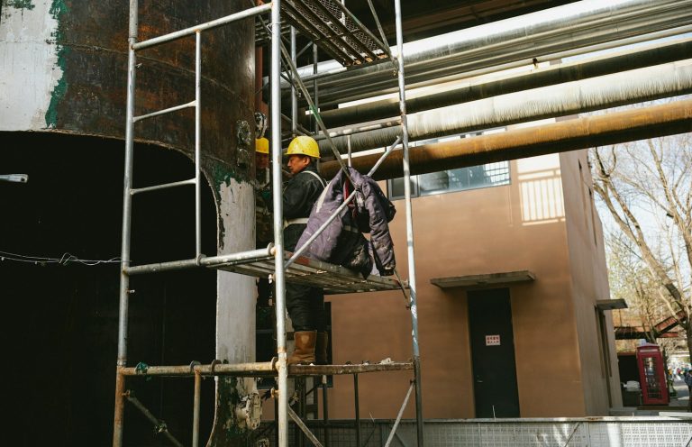 Working at Height Worker in a hard hat climbing a scaffolding beside large pipes.