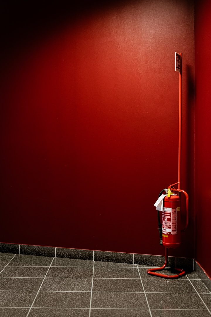 Red fire extinguisher mounted on a wall in a dimly lit corner with dark tiles.